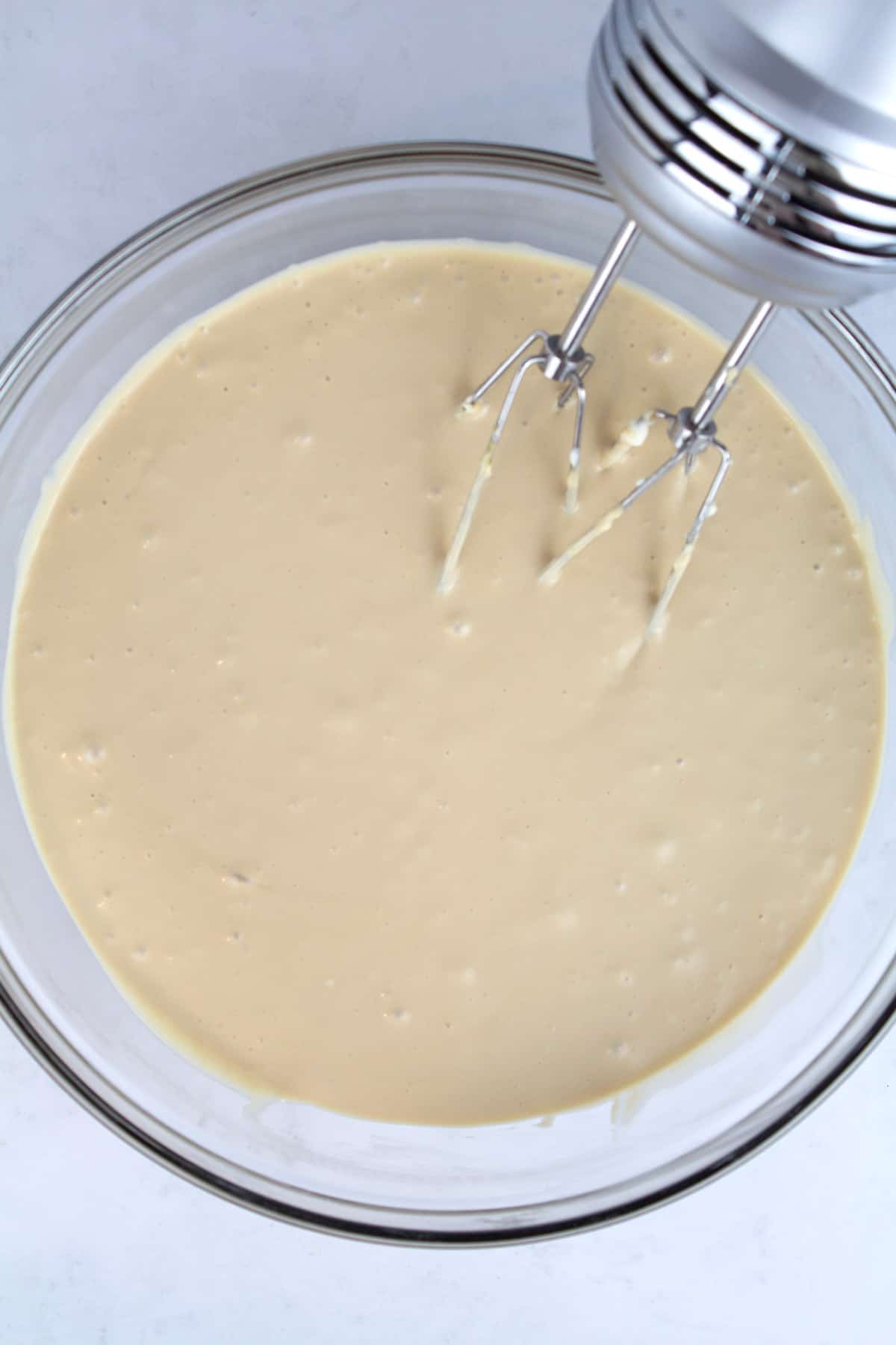 An overhead view of cheesecake batter in a clear glass bowl with a hand mixer resting in the bowl.