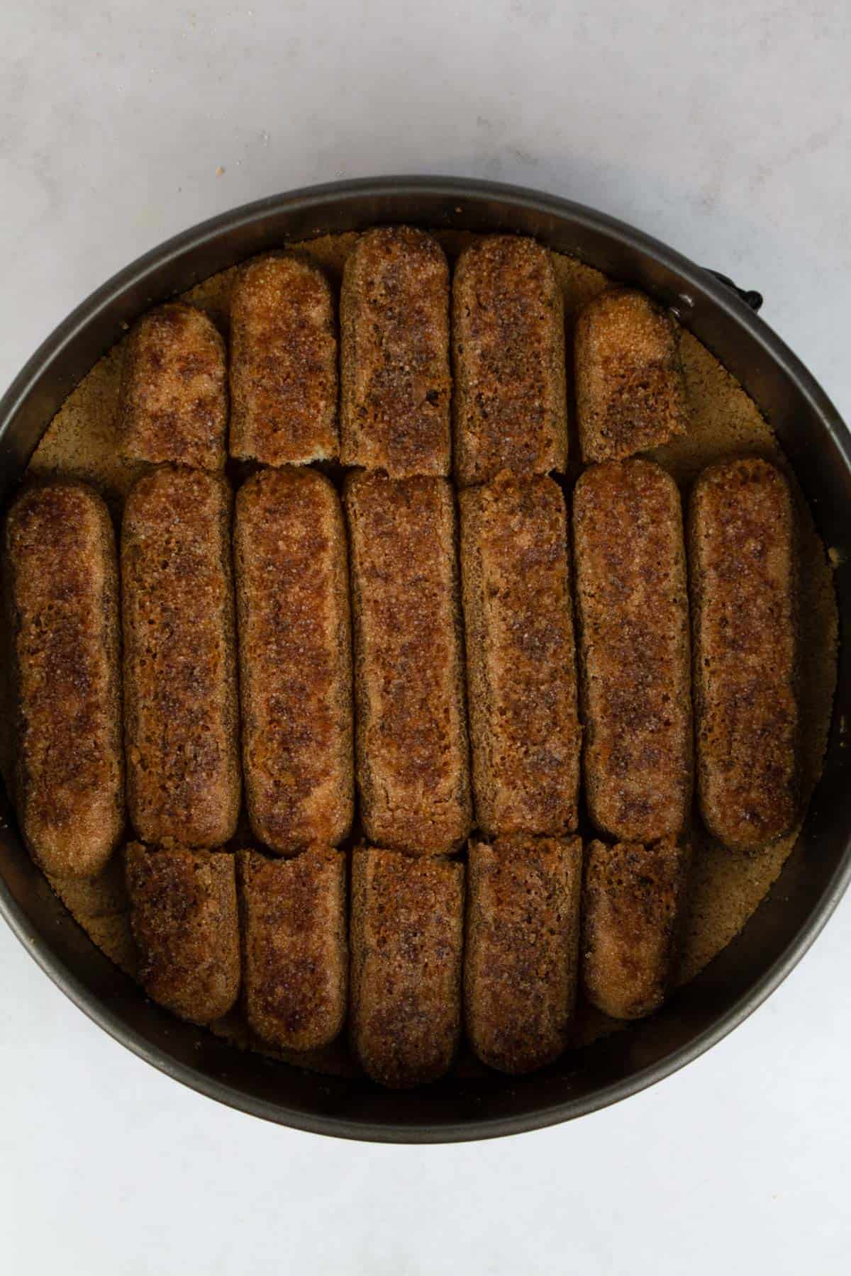 An overhead view of soaked ladyfingers arranged on top of the cheesecake crust.