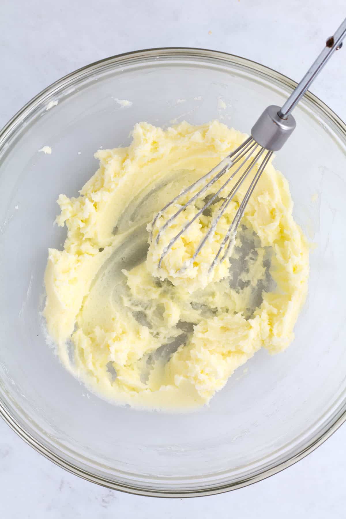 An overhead view of mascarpone and sugar blended together in a clear glass bowl with a whisk.