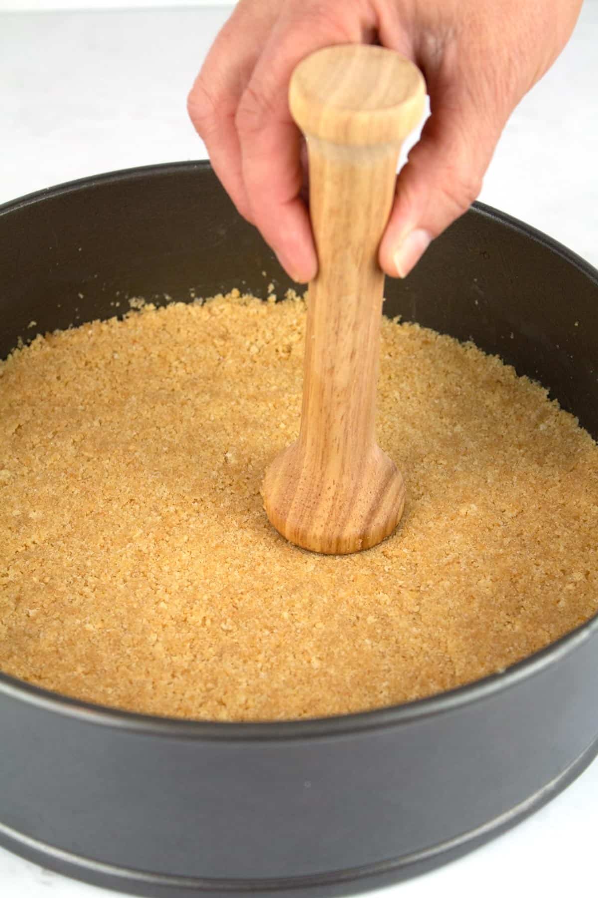 A hand using a tamper to press the crust into a springform pan.