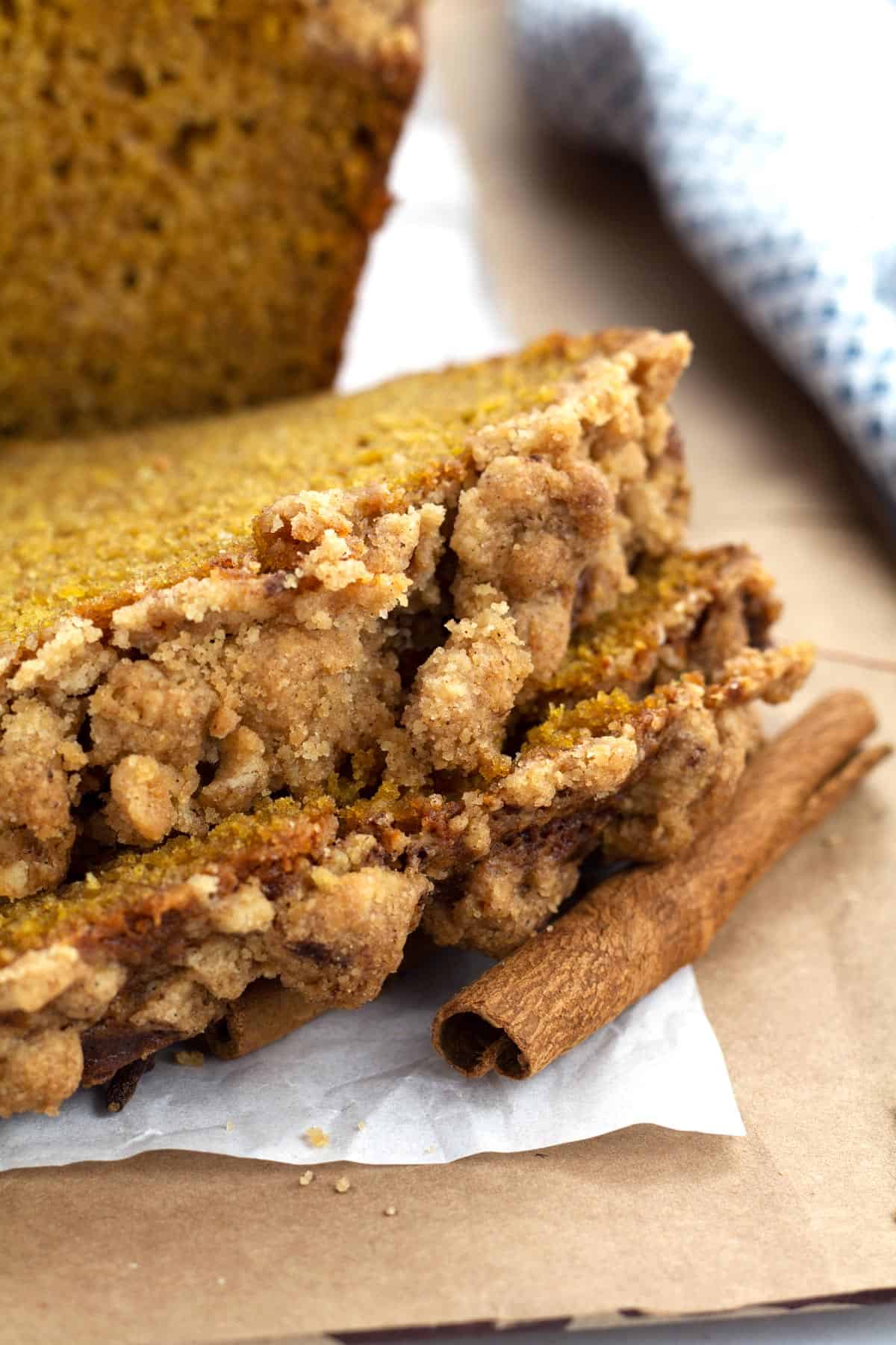 Up close view of two slices of pumpkin bread with moist crumble topping leaning on a cinnamon stick and parchment paper.