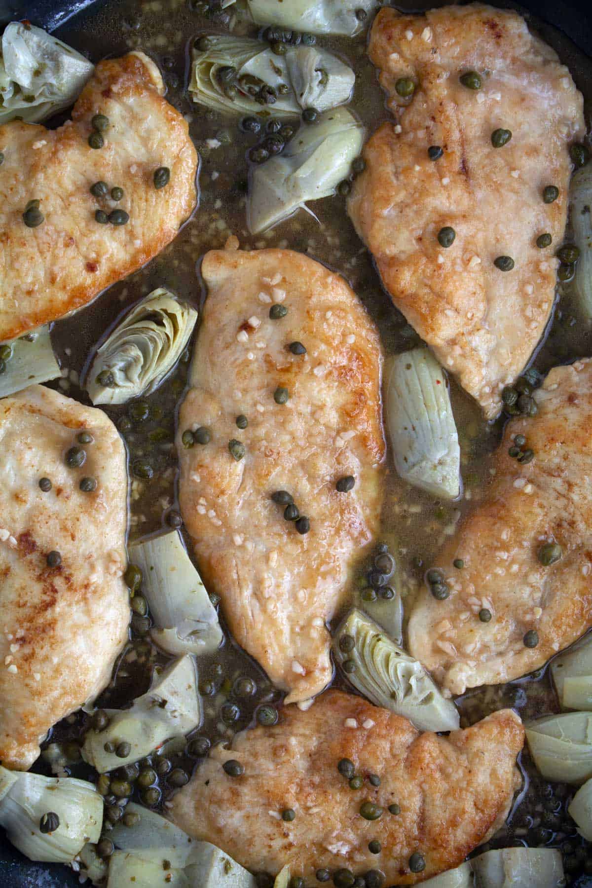 Overhead of chicken pieces cooked in a non-stick skillet with capers and artichoke hearts.