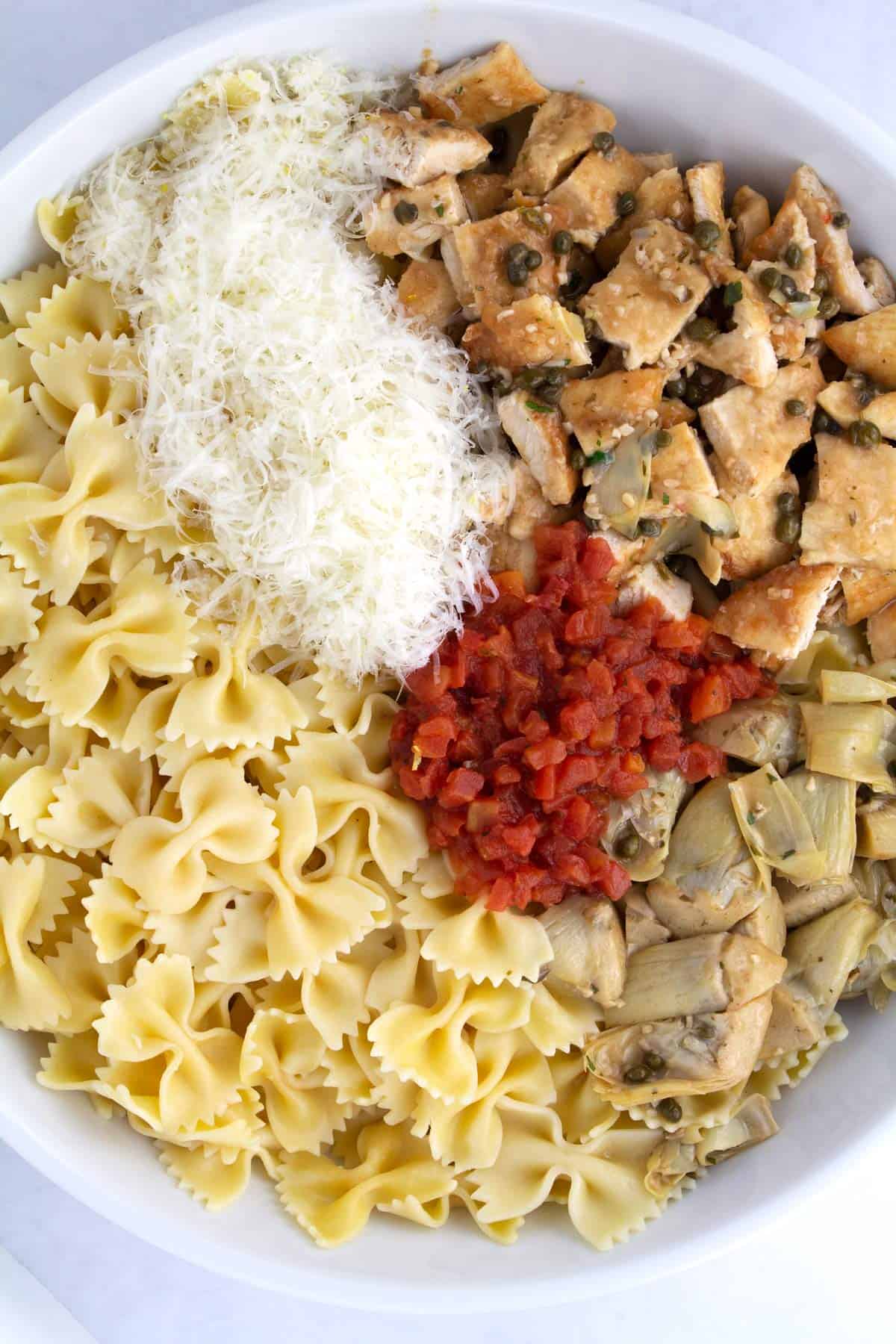 Cooked pasta, tomatoes, parmesa, and chicken piccata in a large white bowl unmixed.