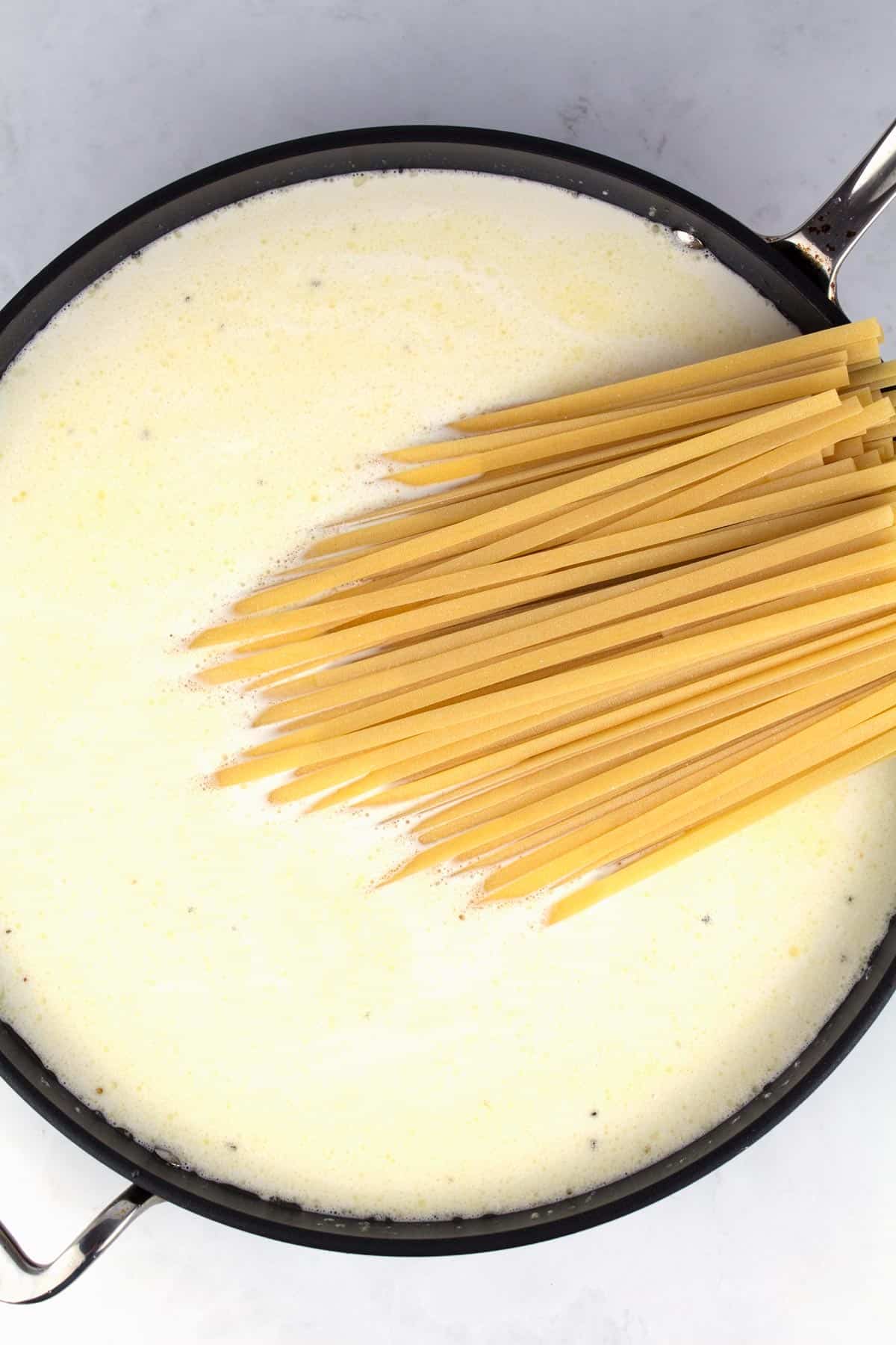 Raw fettuccine noodles sticking out of a skillet filled with cream.