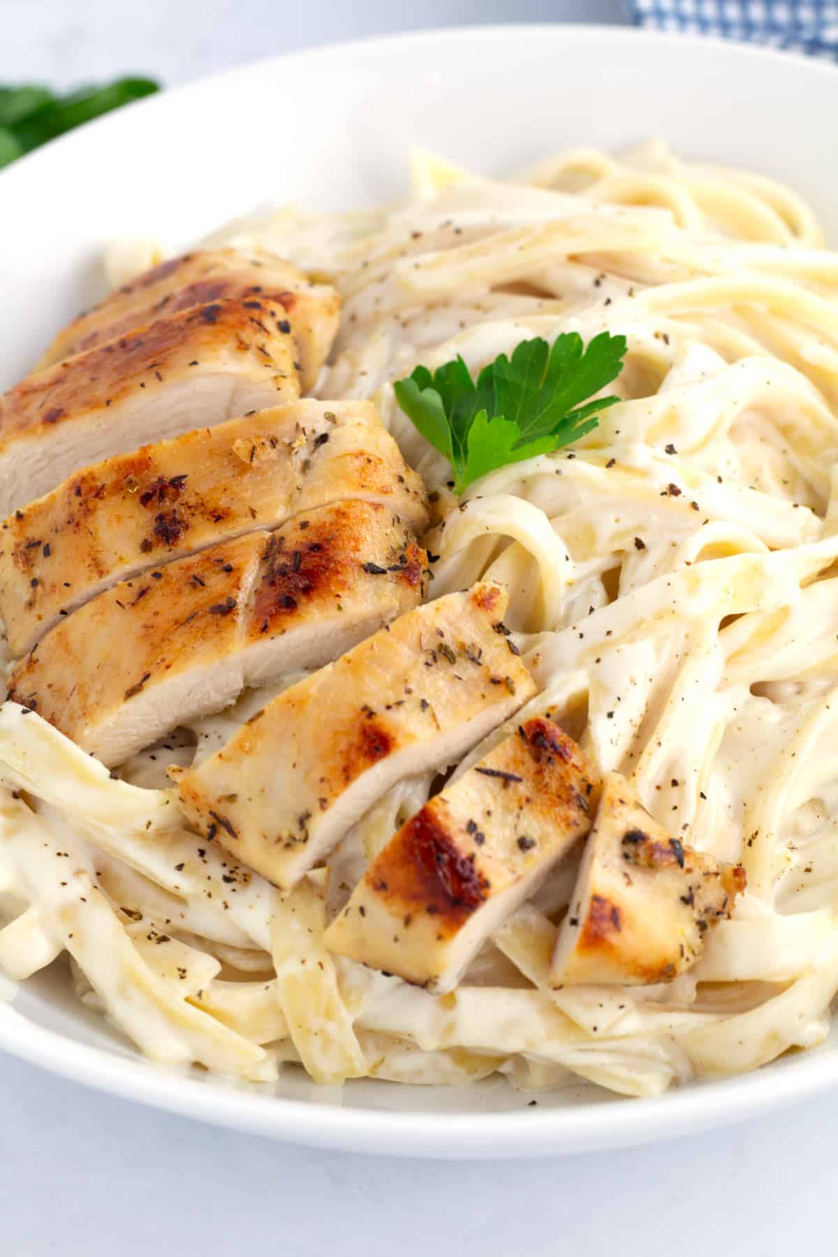 Close up view of sliced chicken on top of fettuccine Alfredo with black pepper and fresh parsley sprinkled on top.