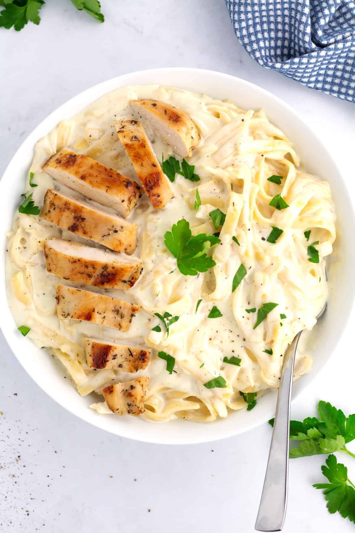 White bowl of chicken Alfredo with golden brown sliced chicken and chopped parsley on top, with a blue checkered towel in the top right corner.