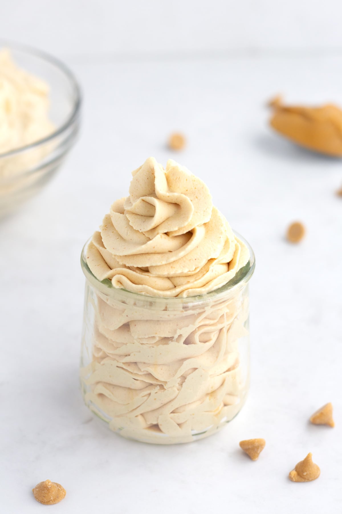 Peanut butter whipped cream piped into a glass jar with peanut butter chips, a spoon of peanut butter, and bowl of whipped cream in the background.