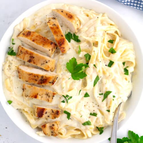 White bowl of chicken fettuccine Alfredo with golden brown sliced chicken and chopped parsley on top.