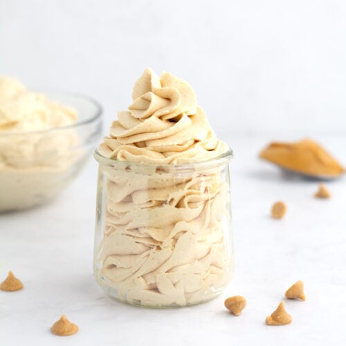 Peanut butter whipped cream piped into a glass jar with peanut butter chips, a spoon of peanut butter, and bowl of whipped cream in the background.
