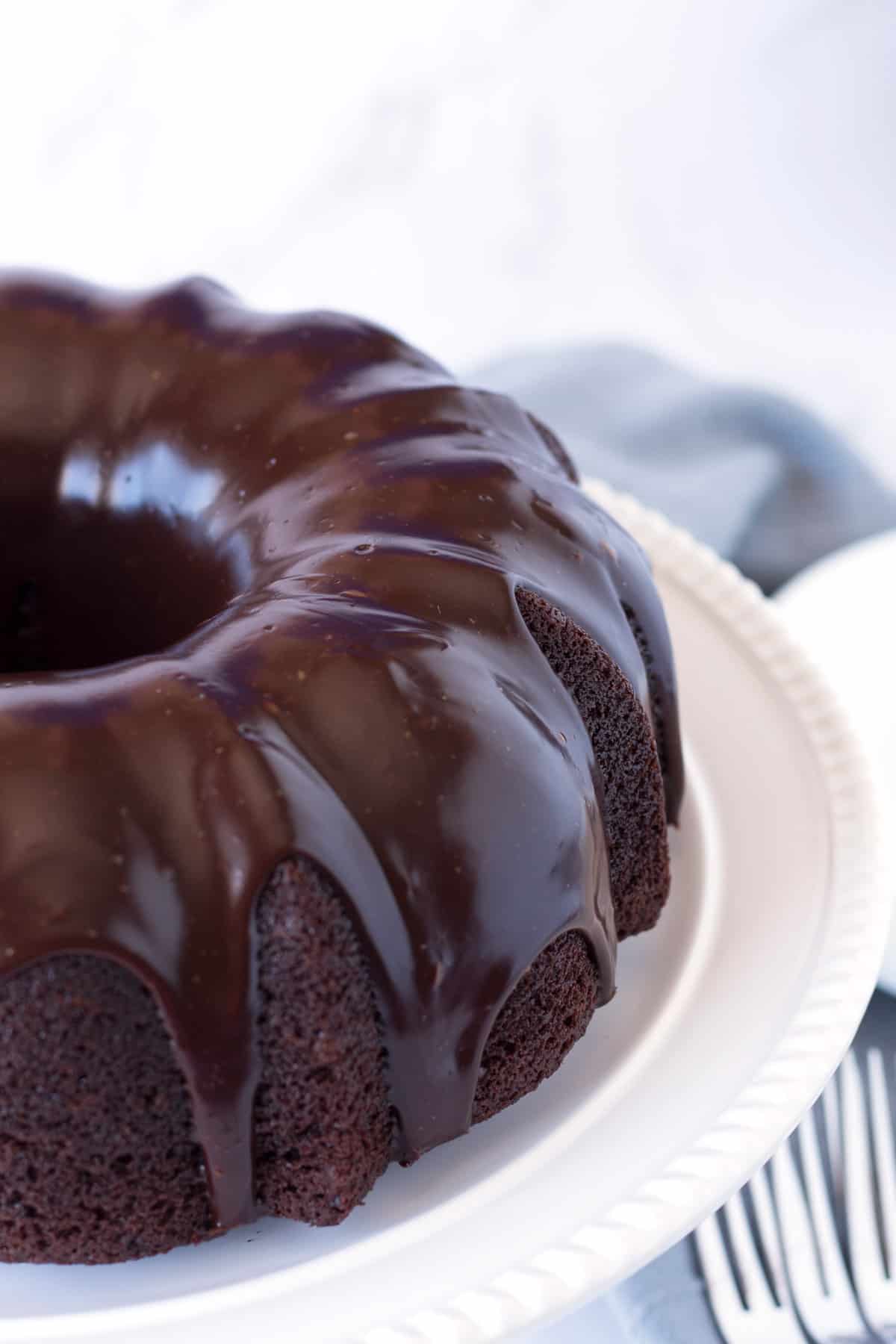 Chocolate bundt cake with chocolate glaze on a white cake stand with a light blue towel in the background.