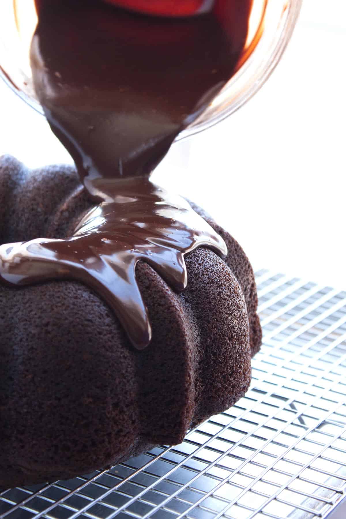 Pouring chocolate ganache out of a glass measuring cup onto a chocolate bundt cake.