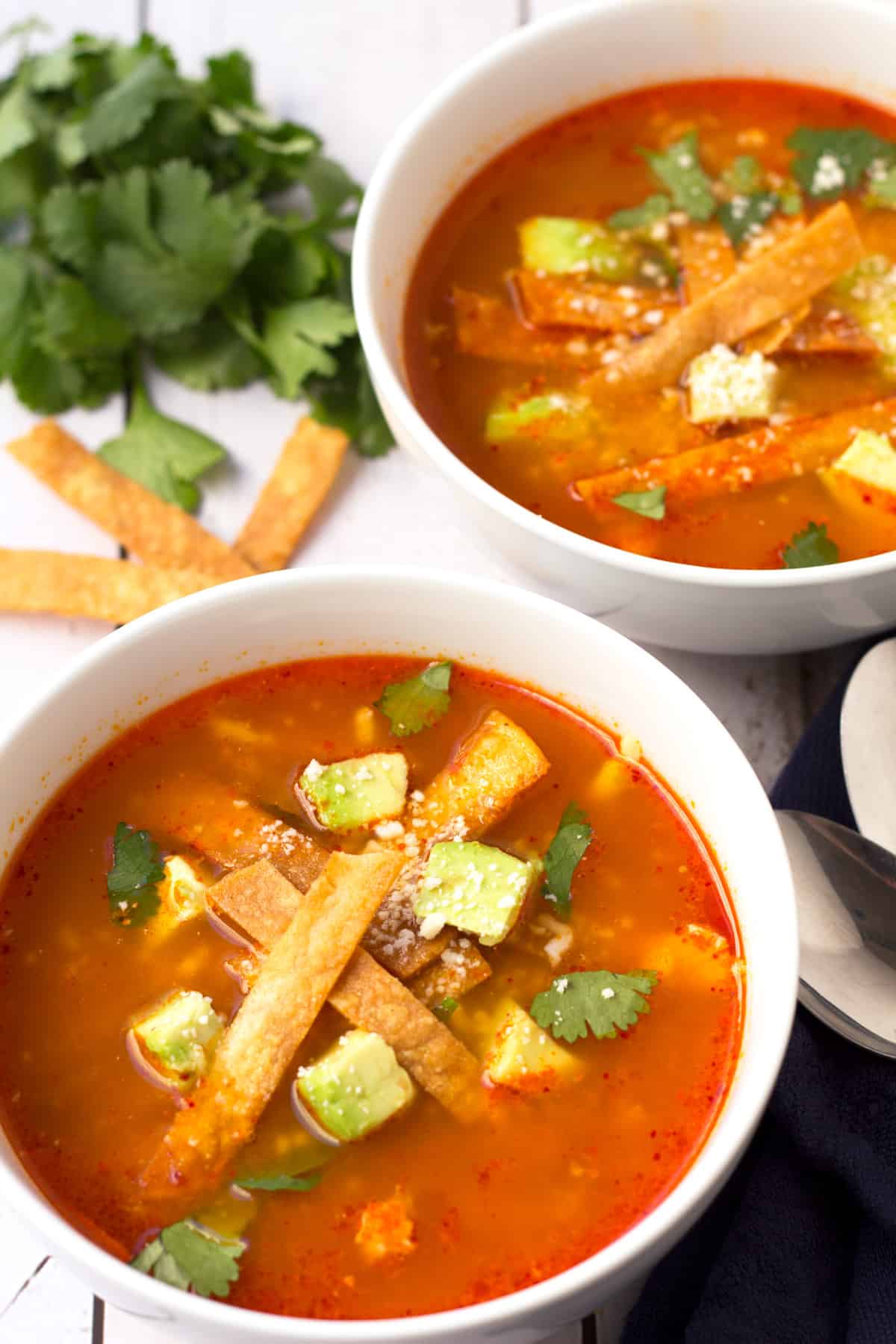 Two bowls of chicken tortilla soup with thin tortilla strips and avocado on top.
