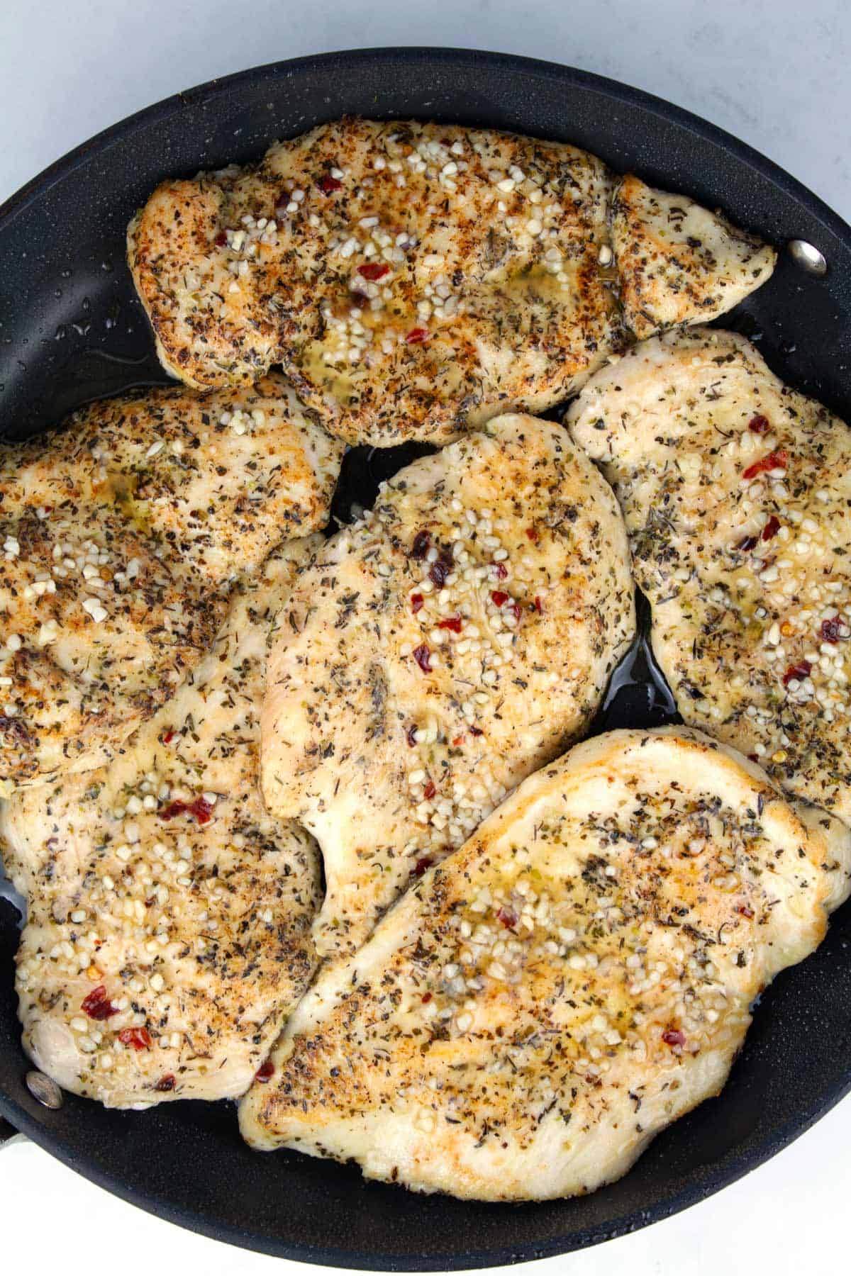 Overhead of skillet with six thin chicken breasts seared and covered with garlic butter.