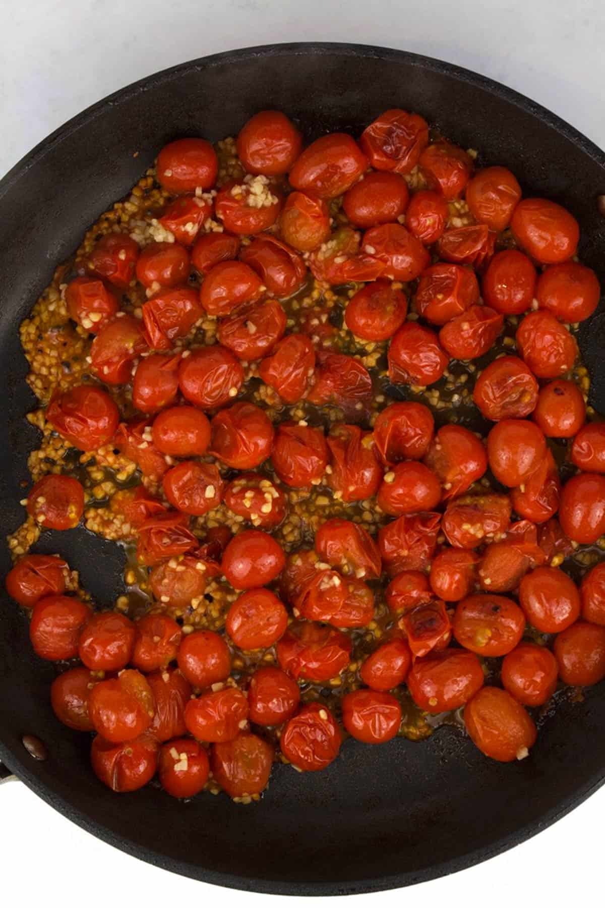 A black skillet with burst, roasted tomatoes and minced garlic.