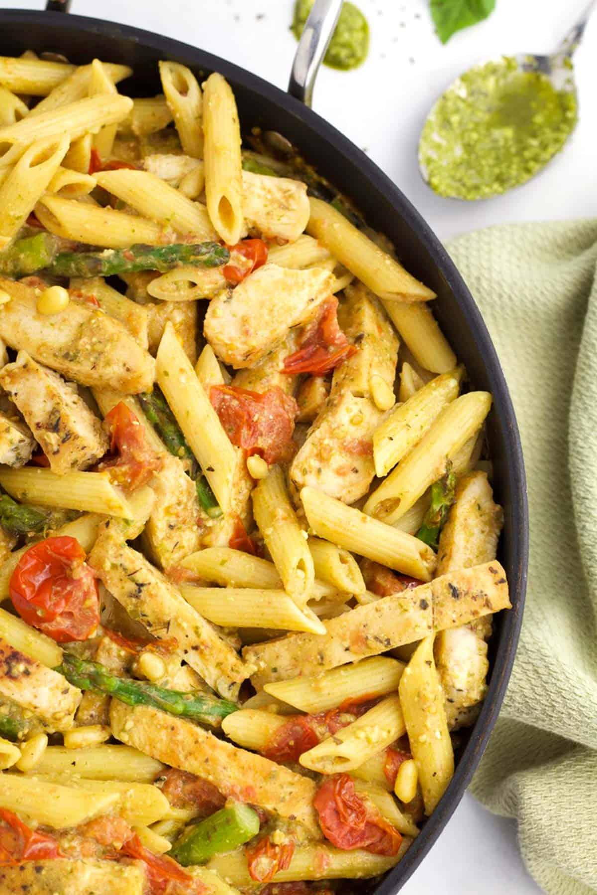 A black skillet full of chicken pesto pasta with tomatoes and veggies, with a green towel on the side of the pan.