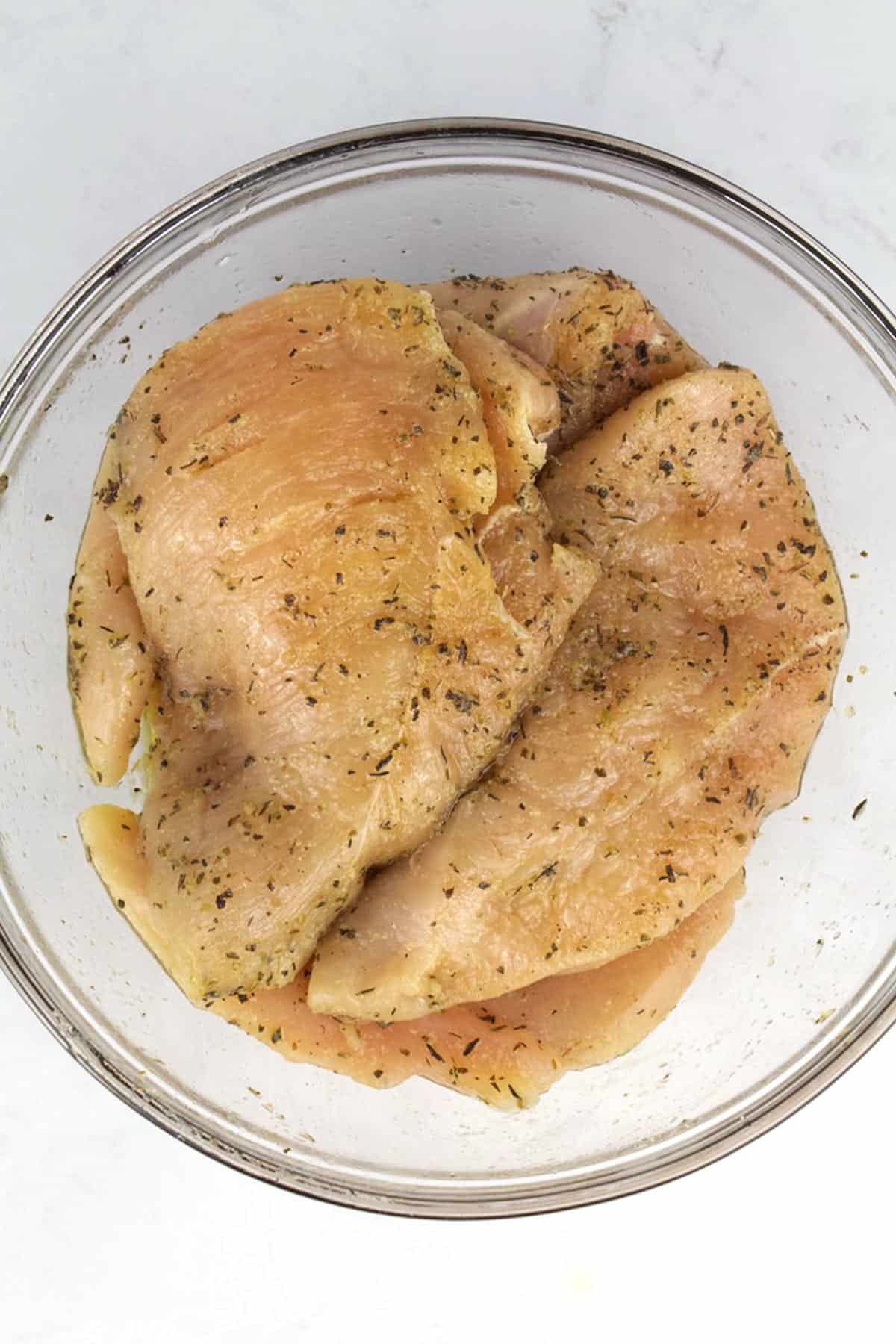 A glass bowl with raw chicken breasts seasoned with dried herbs and olive oil.