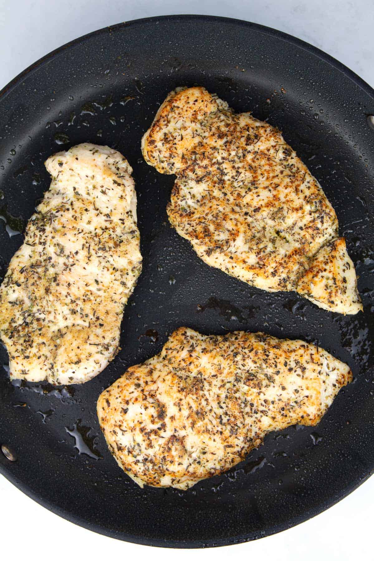 Overhead view of a black skillet with three thin chicken breasts searing.