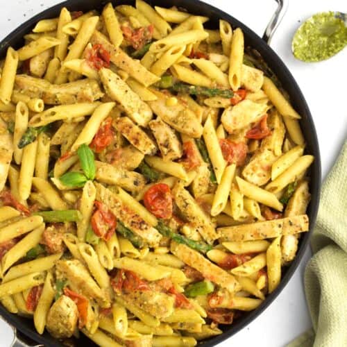A black skillet full of chicken pesto pasta with tomatoes and veggies, with a green towel on the side of the pan.