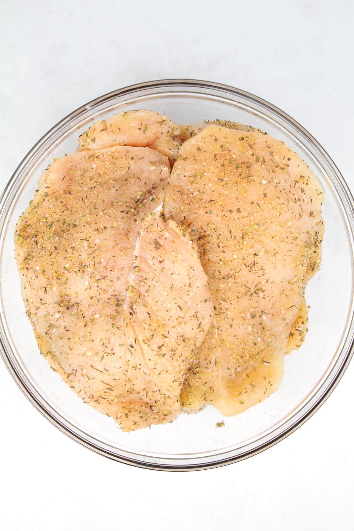 Clear glass bowl with raw chicken breasts sprinkled with seasonings.