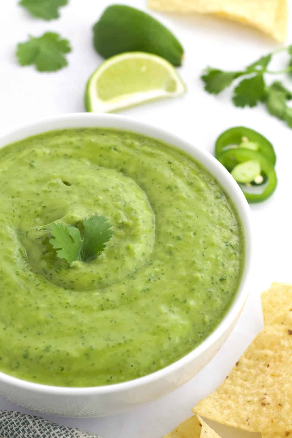 White bowl of green salsa with cilantro, chips, limes, and jalpeno slices in the background.