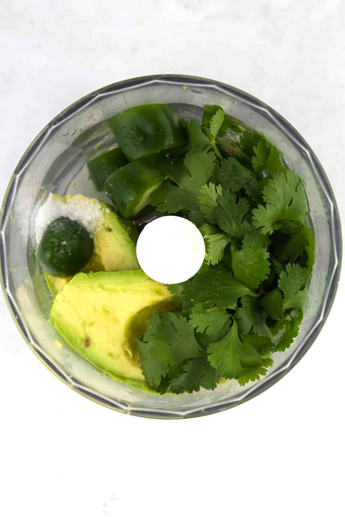 Food processor with chunks of avocado, jalapeno, cilantro, and salt in the middle.