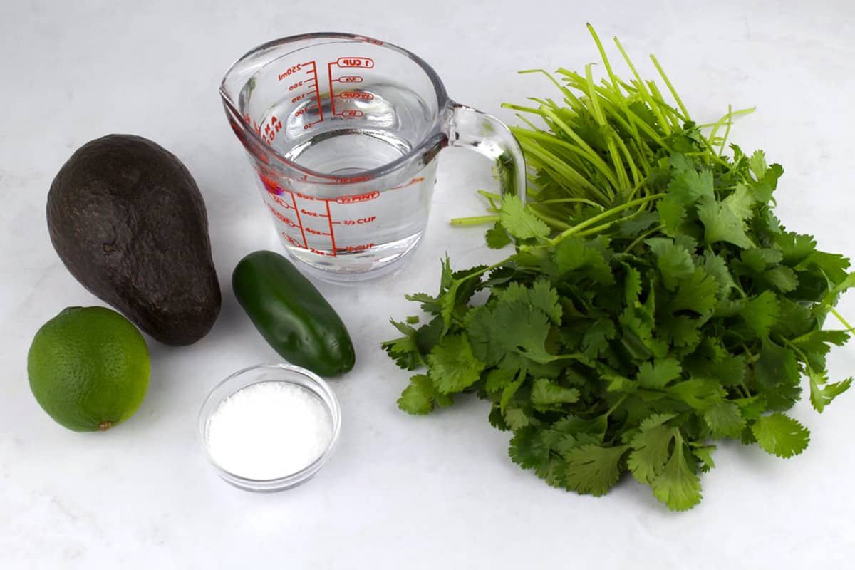 Ingredients for salsa on a white counter, including cilantro, jalapeno, avocado, salt, and lime.