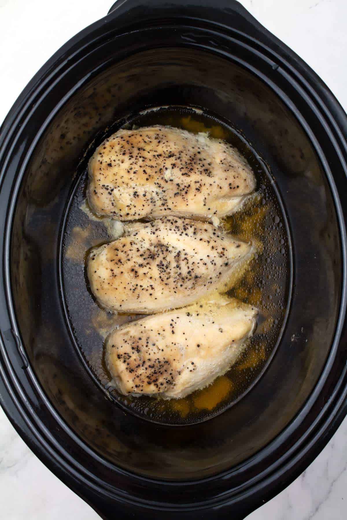 Overhead view of 3 cooked chicken breasts and braising liquid in a black slow cooker.