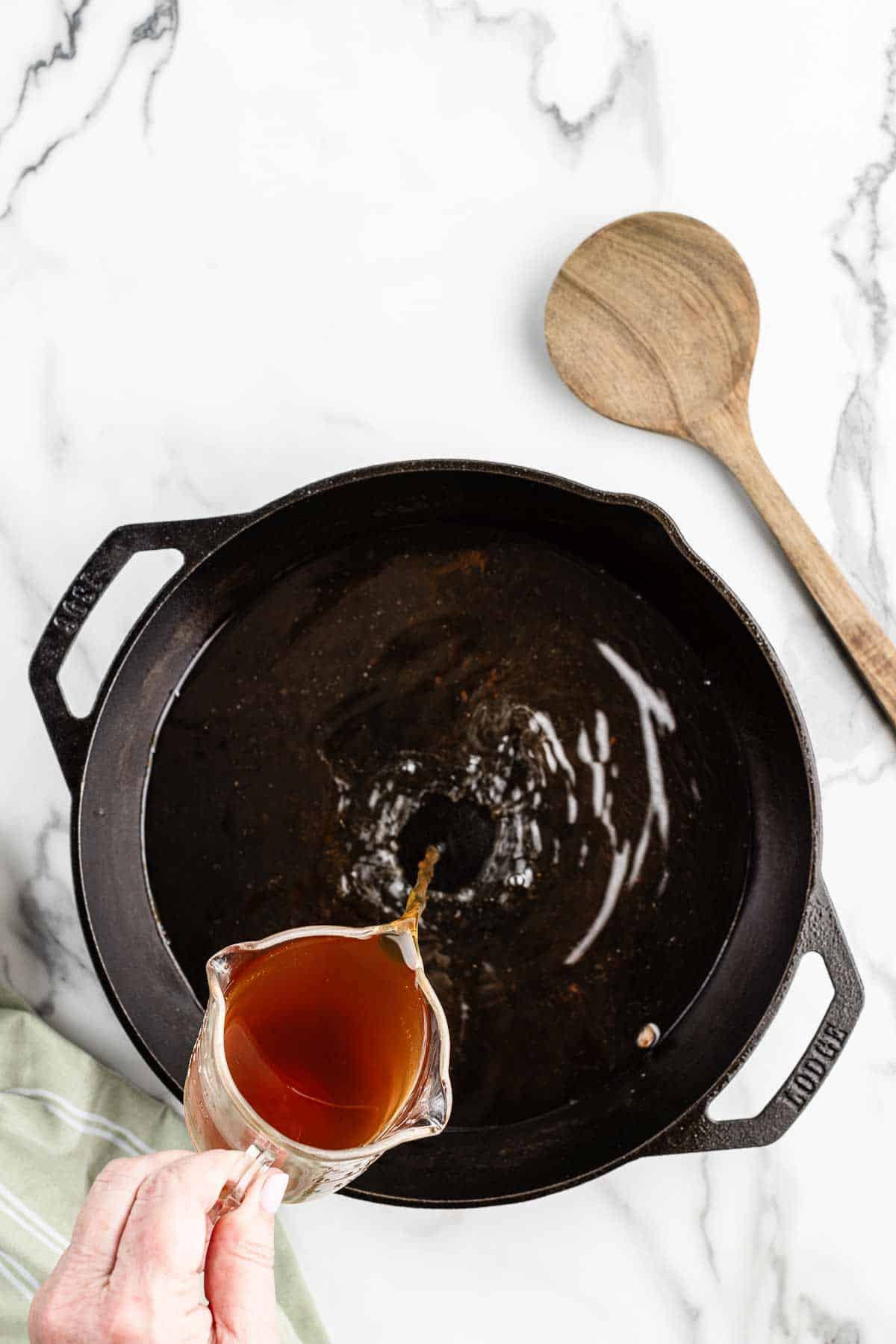 Overhead view of a hand pouring beef broth into an empty black skillet.