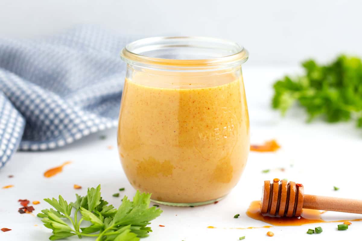 A horizontal photo of a jar of hot honey mustard with a honey stick on the right, a blue towel in the background on the left, and bunches of green parsley throughout the scene.