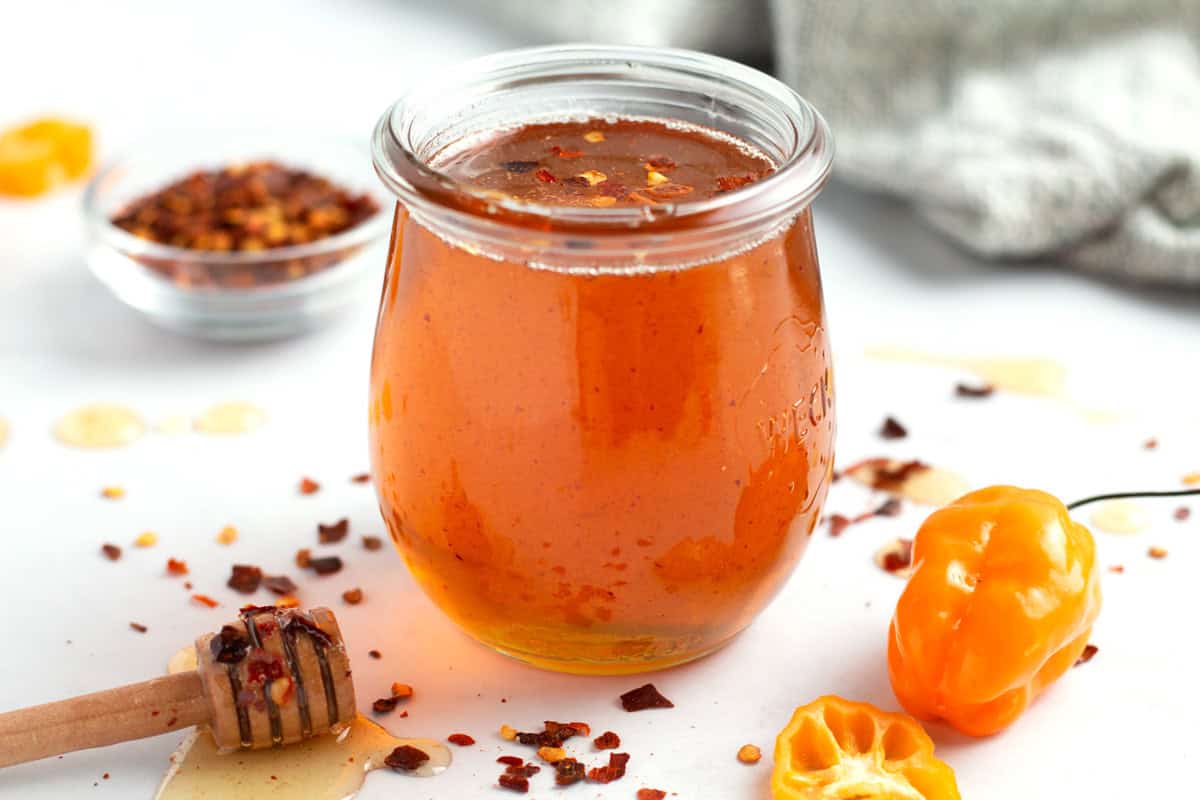 A jar of spicy honey on a white counter with a honey stick, drips of honey, red pepper flakes, and a habanero pepper sitting around the jar.