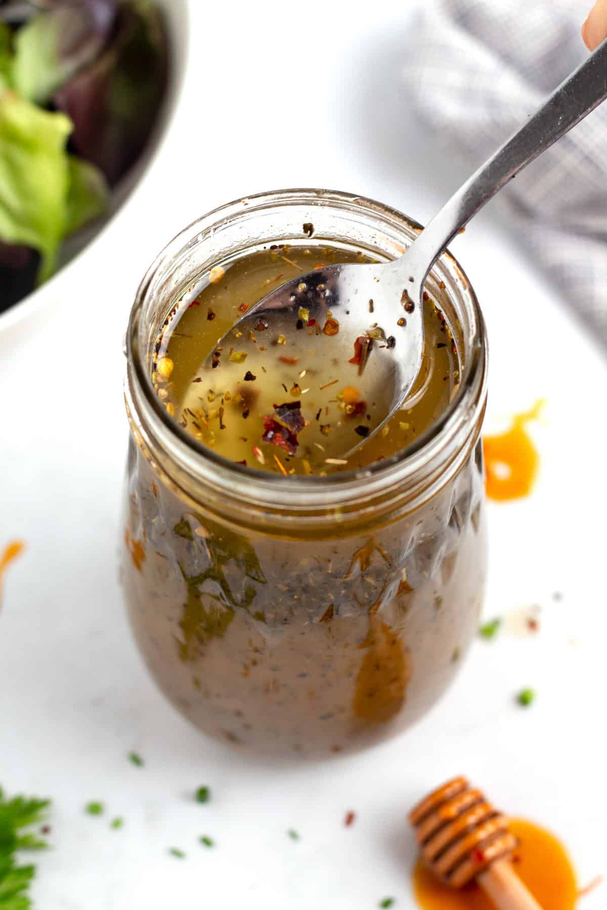 Jar of vinaigrette with a spoon being lifted out of it and a bowl of green lettuce in the background.