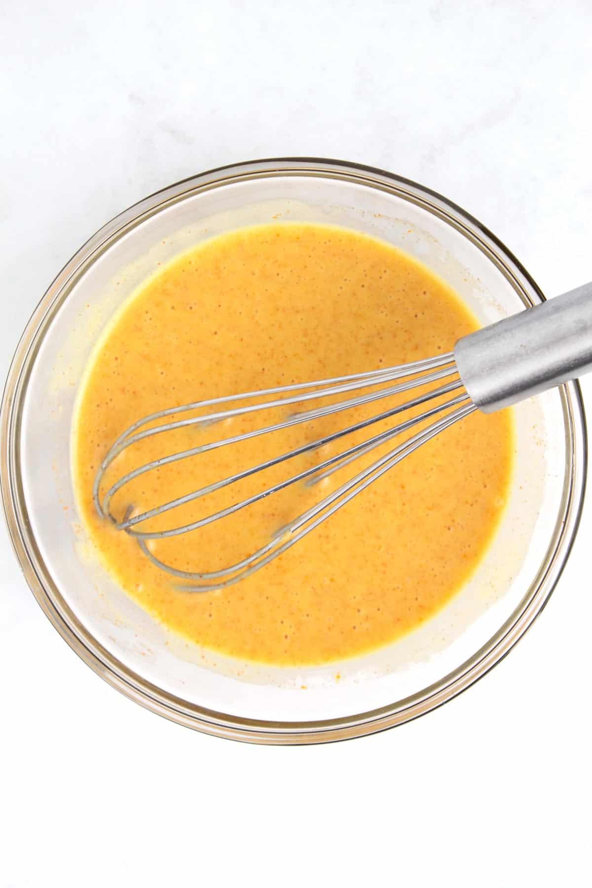An overhead view of a clear glass bowl of combined hot honey mustard ingredients with a whisk sitting in the bowl.