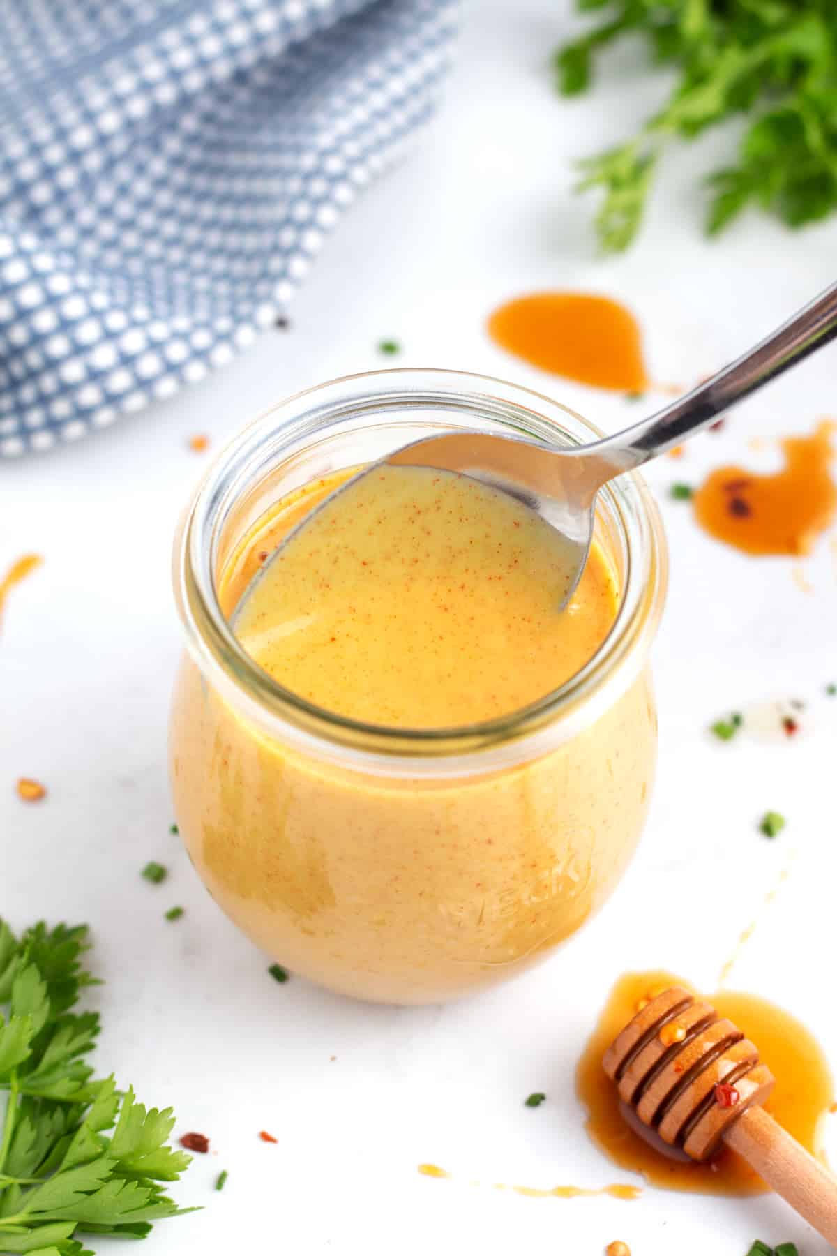 A silver spoon coming out of a jar of hot honey mustard surrounded by a honey stick, parsley, chives, and red pepper flakes on the counter.