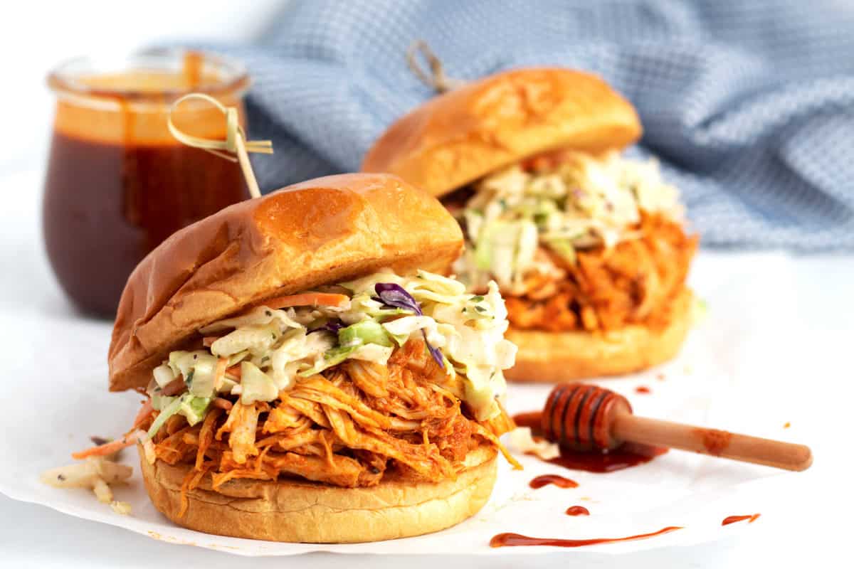 Two shredded chicken sandwiches on a piece of parchment paper with red honey sauce drips, a jar of honey sauce and a light blue towel throughout the scene.