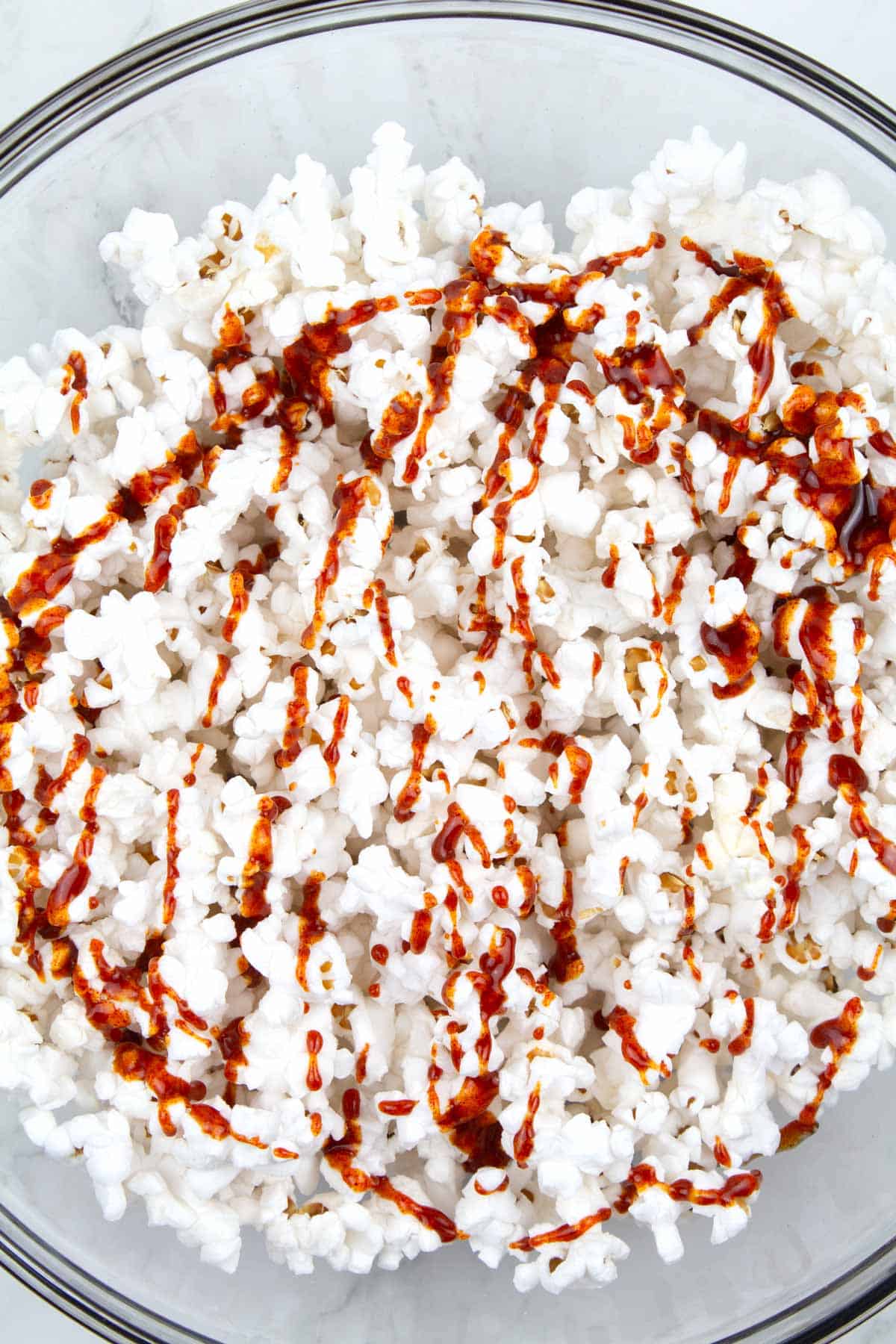 Popped popcorn in a bowl drizzled with red hot honey sauce before mixing together.