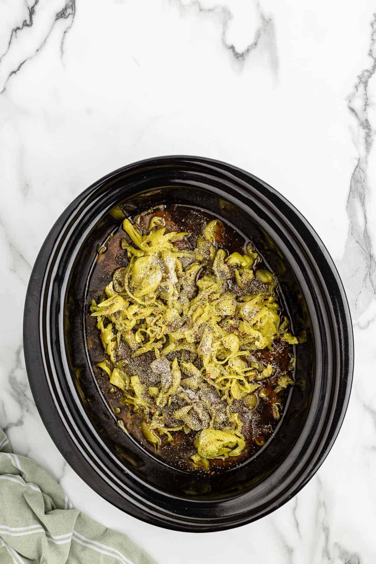 Overhead of slow cooker with seasonings and peppers sprinkled on top of the meat.