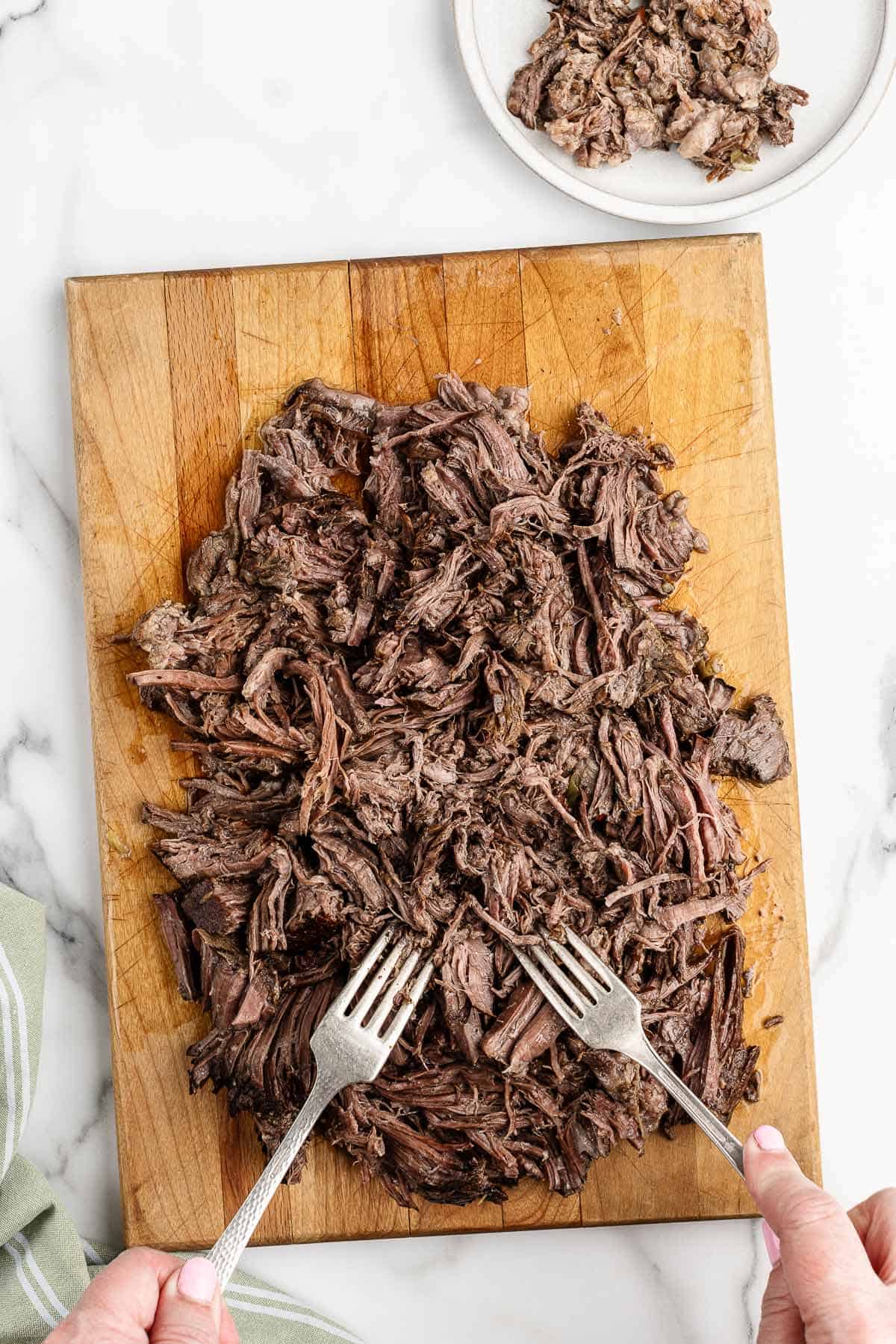 Overhead view of two hands shredding beef with forks on a wooden cutting board.