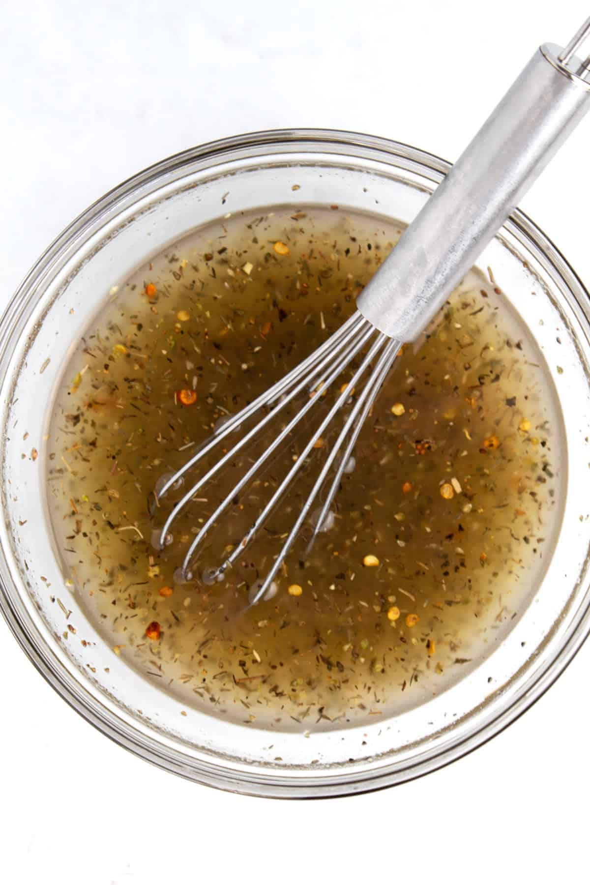 Overhead view of a clear glass bowl with an herby vinaigrette dressing and a whisk in the bowl.
