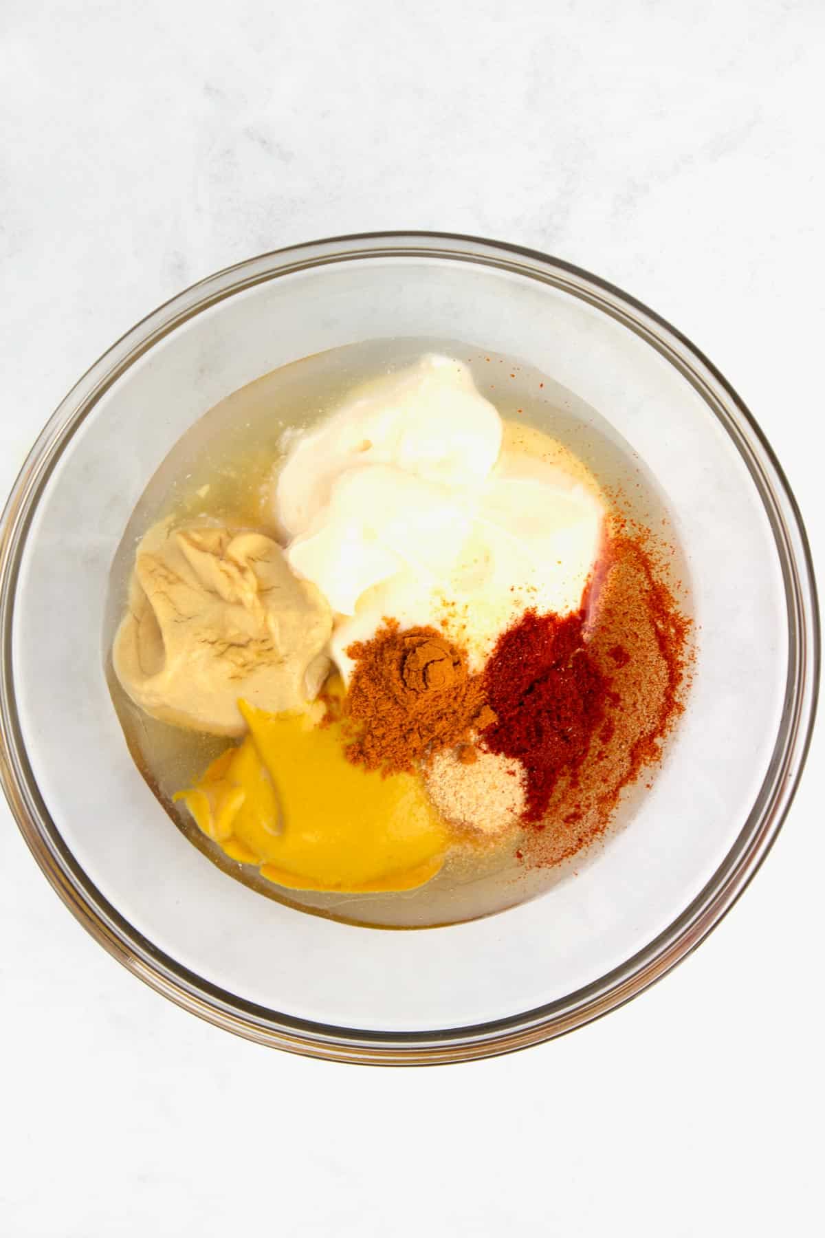 An overhead view of a clear glass bowl with piles of mayo, mustard, lemon juice, honey, and spices unmixed in the bowl.