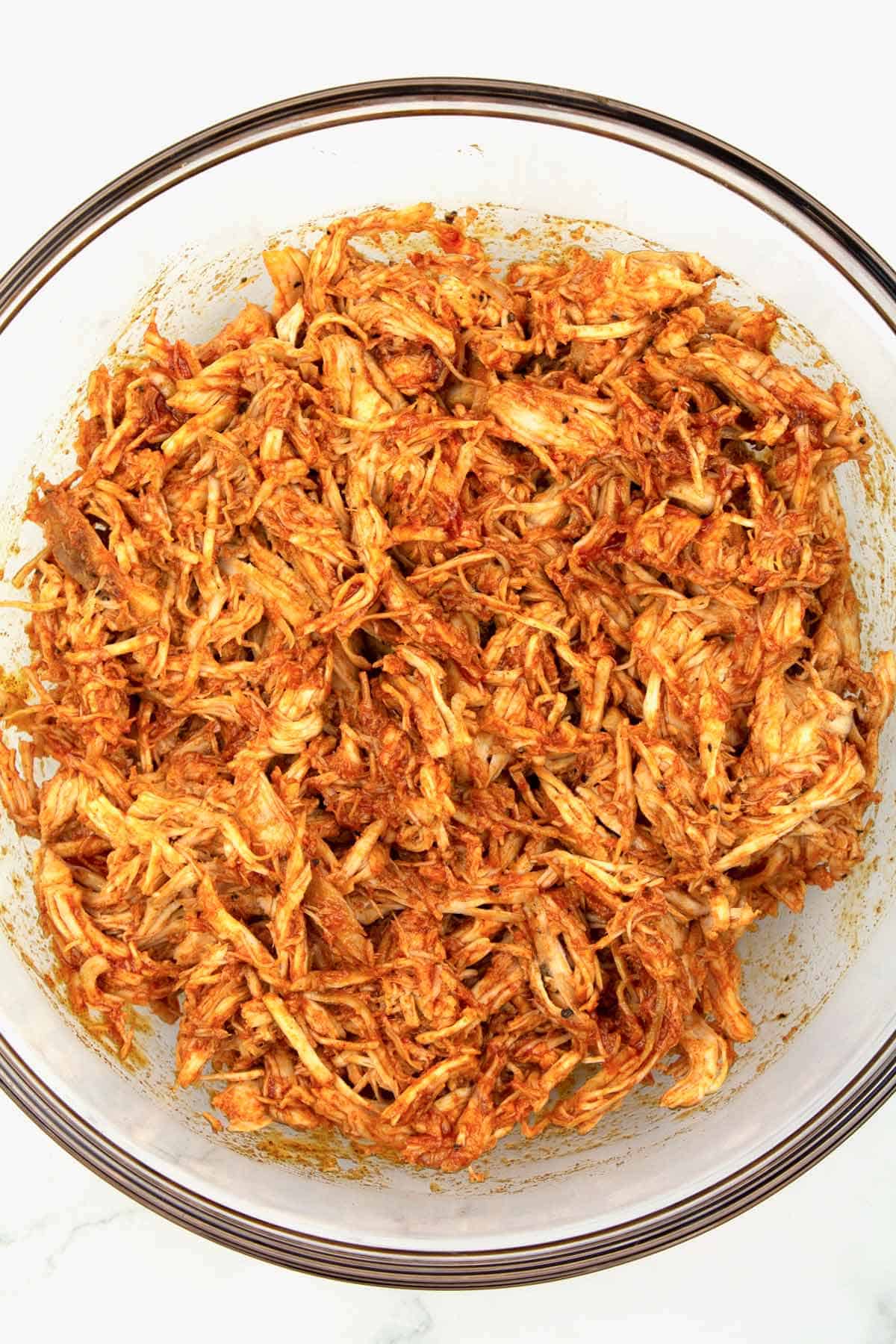 Overhead view of a clear glass bowl full of shredded chicken covered in a red spicy honey sauce.