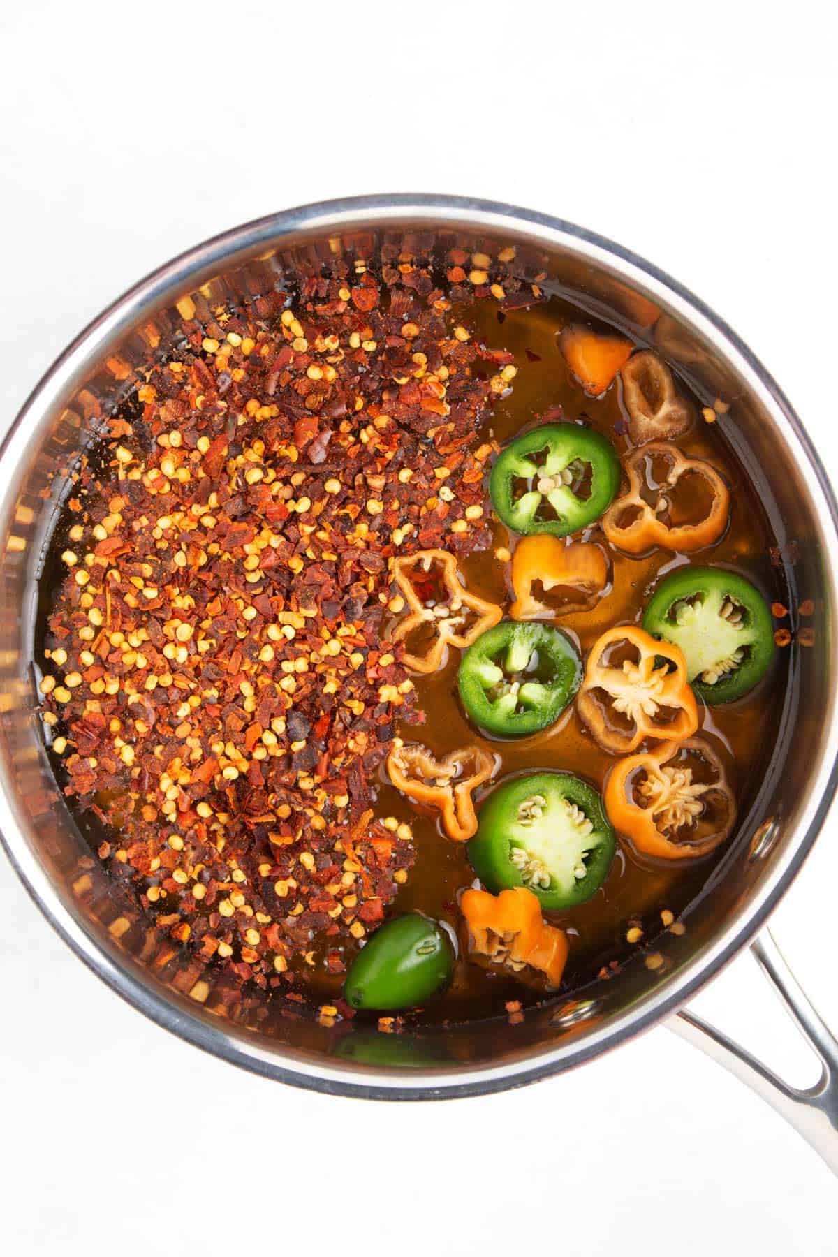 Overhead view of a silver saucepan with honey, pepper flakes, habaneros, and jalapenos inside.