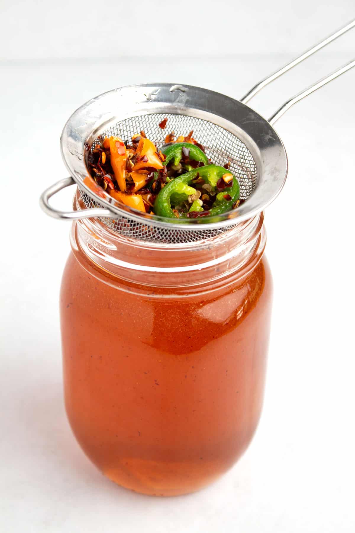 Glass jar full of honey with a strainer on the top full of red pepper flakes and sliced peppers.