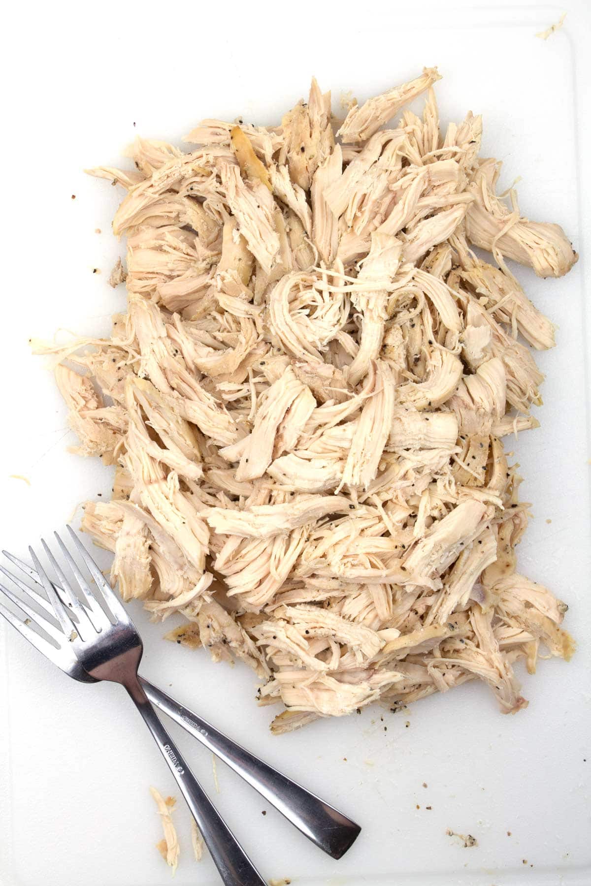 Overhead view of a pile of shredded chicken on a white cutting board with two forks sitting next to the chicken.