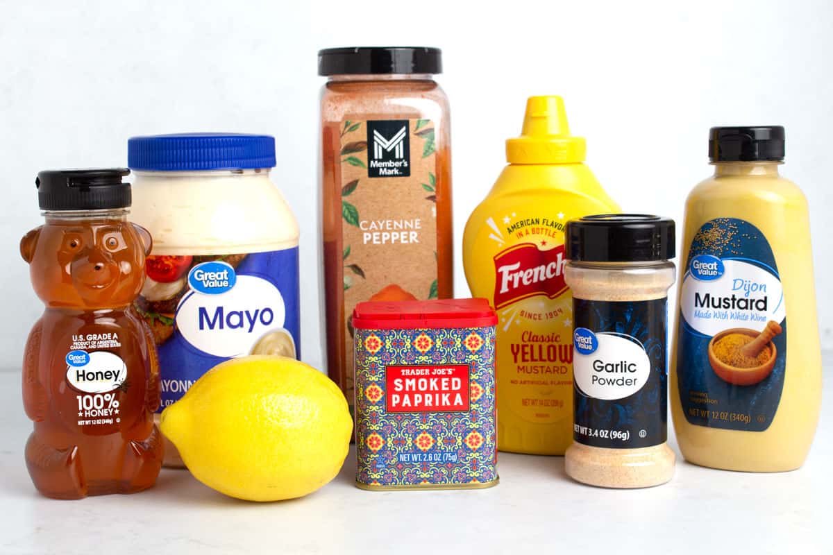 Bottles of honey, mayo, mustards, spices, and a lemon on a white countertop.