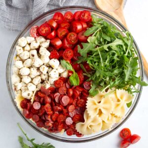 Pepperoni, bowtie pasta, arugula, cherry tomatoes, mozzarella pearls, and dressing layered in glass dish.