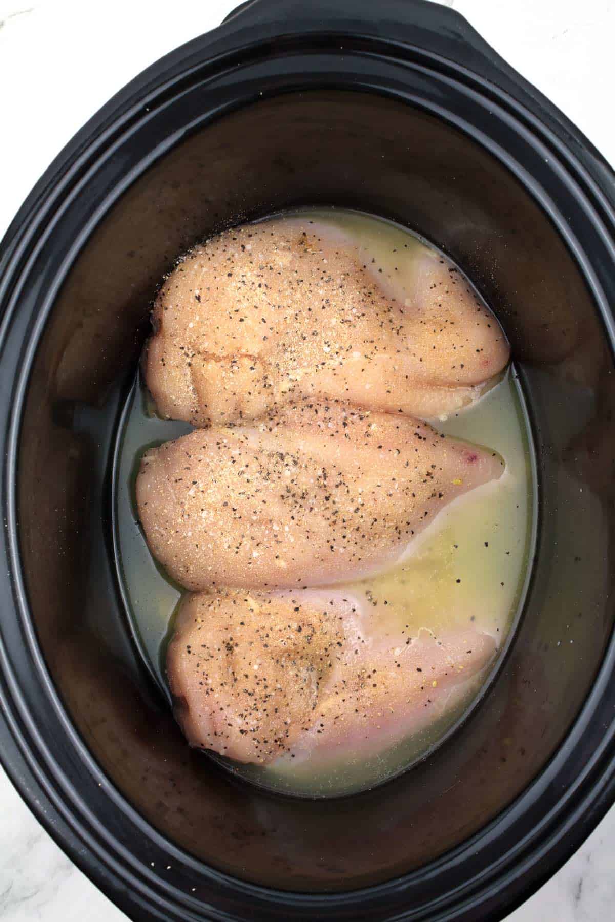 Overhead view of three raw chicken breasts sprinkled with pepper and partially covered in chicken broth in a black crock pot.