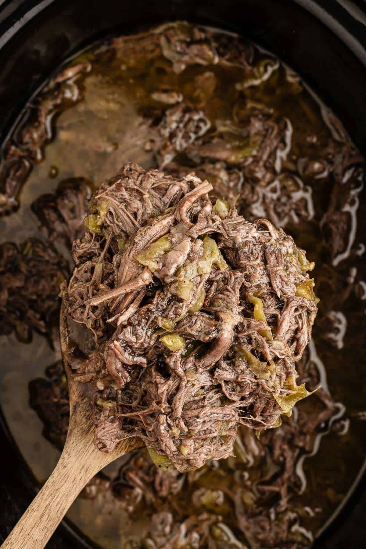 A wooden spoon holding slow cooker pepperoncini beef above a slow cooker.