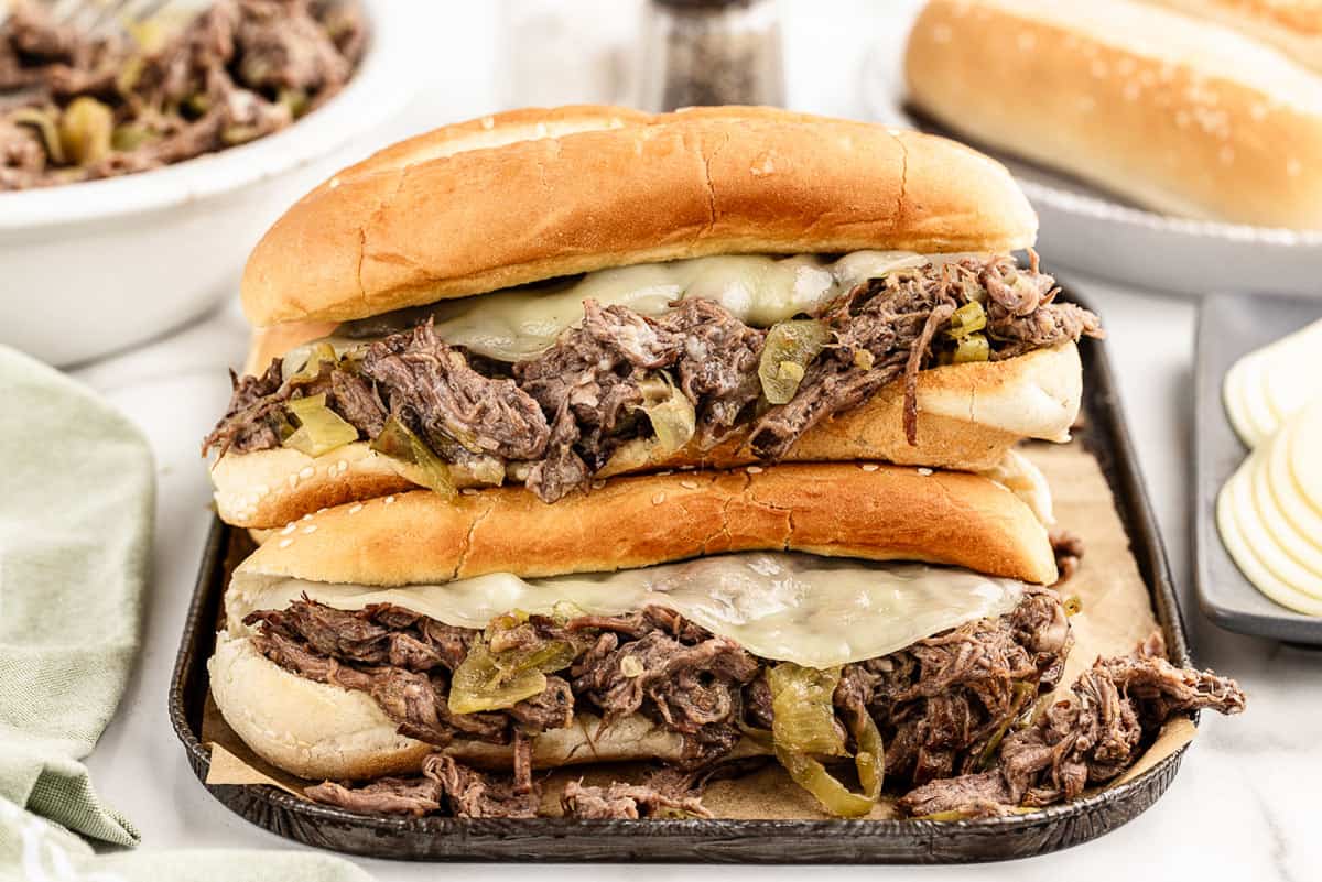 Two Italian beef sandwiches stacked on top of each other on a cookie sheet with extra rolls, meat, and cheese in the background.