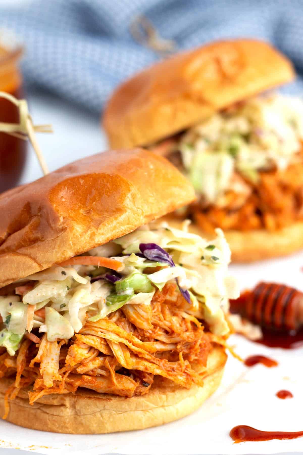 A chicken sandwich made with hot honey shredded chicken, coleslaw, and a hamburger bun with honey drizzles and another sandwich in the background.