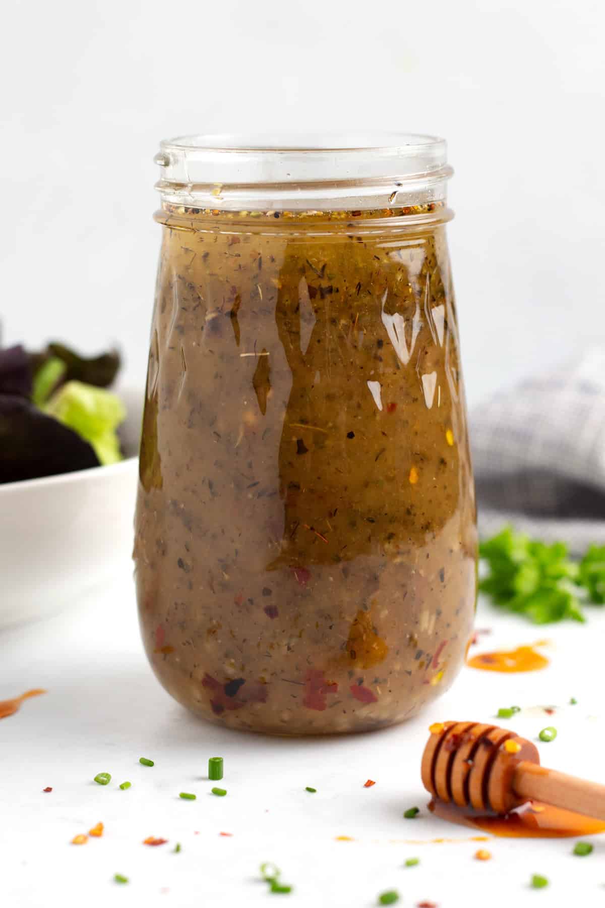 A close up view of a jar of salad dressing with fresh herbs sprinkled around the jar, a honey stick laying in front of the jar, and a bowl of lettuce behind the jar.